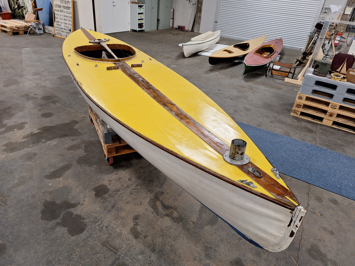 Segelkanot, klass III B, med Güntherrigg och centerbord. Fall, skot och block saknas. Byggd i mahogny, furu och duk. 
Vita fribord och gult däck. Mittfiskar och sarg i mahogny.