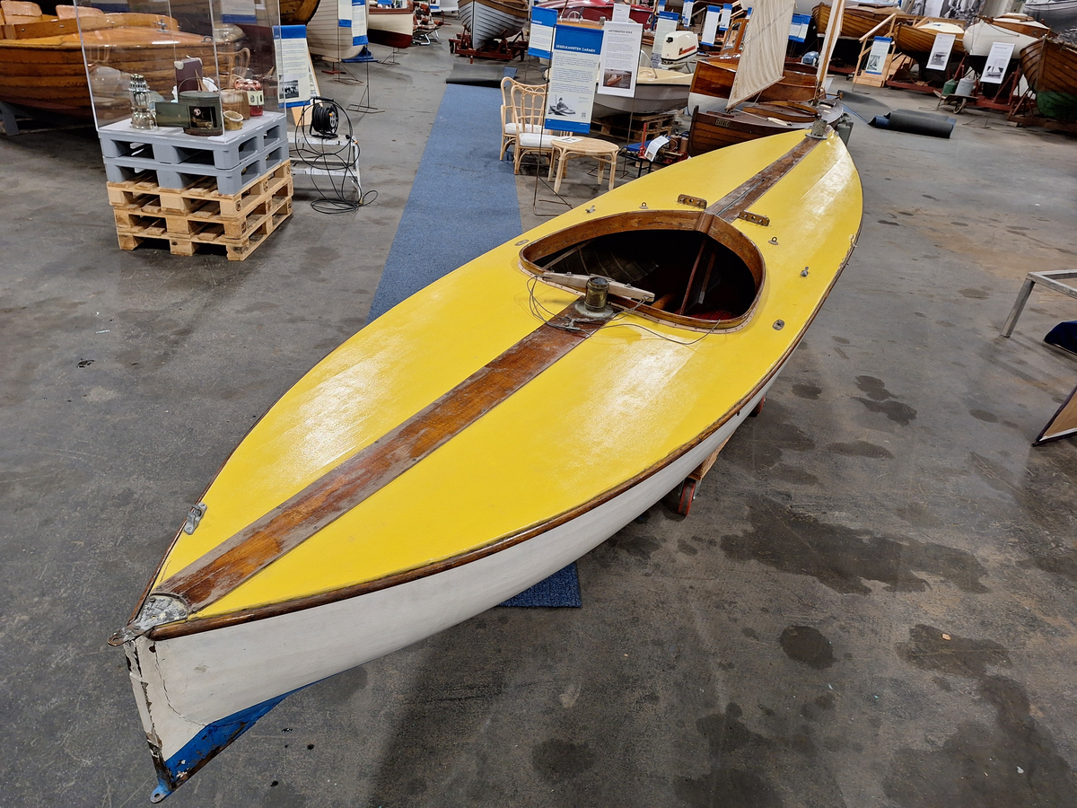 Segelkanot, klass III B, med Güntherrigg och centerbord. Fall, skot och block saknas. Byggd i mahogny, furu och duk. 
Vita fribord och gult däck. Mittfiskar och sarg i mahogny.