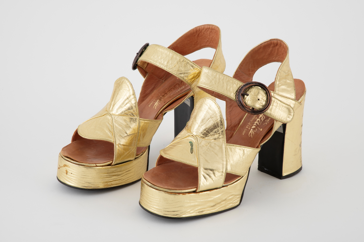 Gullsandaler med kraftig platåsåle og hæl. Åpen hæl og tå med remmer rundt hæl og ankel, den siste med en sirkelrund metallspenne. Brede, kryssende remmer over vristen. Beige skinnfôr.