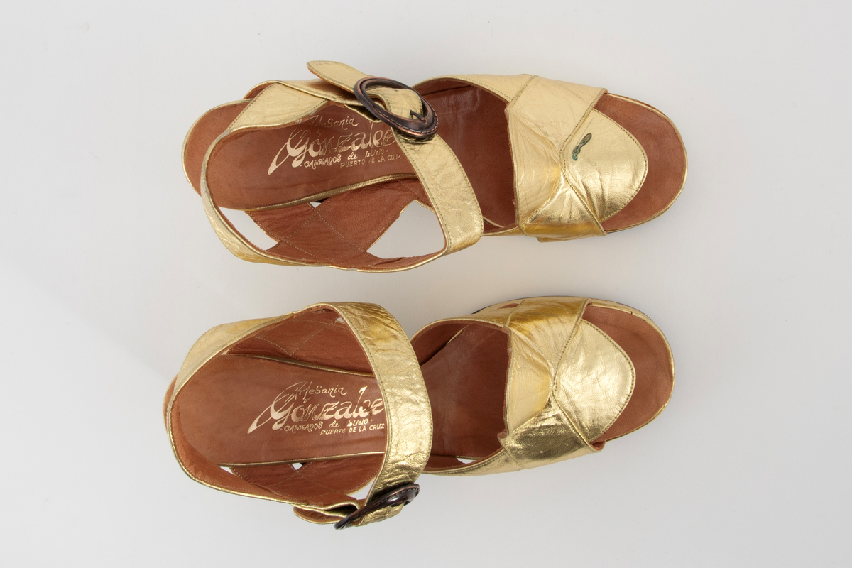 Gullsandaler med kraftig platåsåle og hæl. Åpen hæl og tå med remmer rundt hæl og ankel, den siste med en sirkelrund metallspenne. Brede, kryssende remmer over vristen. Beige skinnfôr.