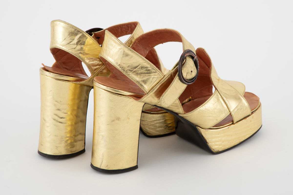 Gullsandaler med kraftig platåsåle og hæl. Åpen hæl og tå med remmer rundt hæl og ankel, den siste med en sirkelrund metallspenne. Brede, kryssende remmer over vristen. Beige skinnfôr.