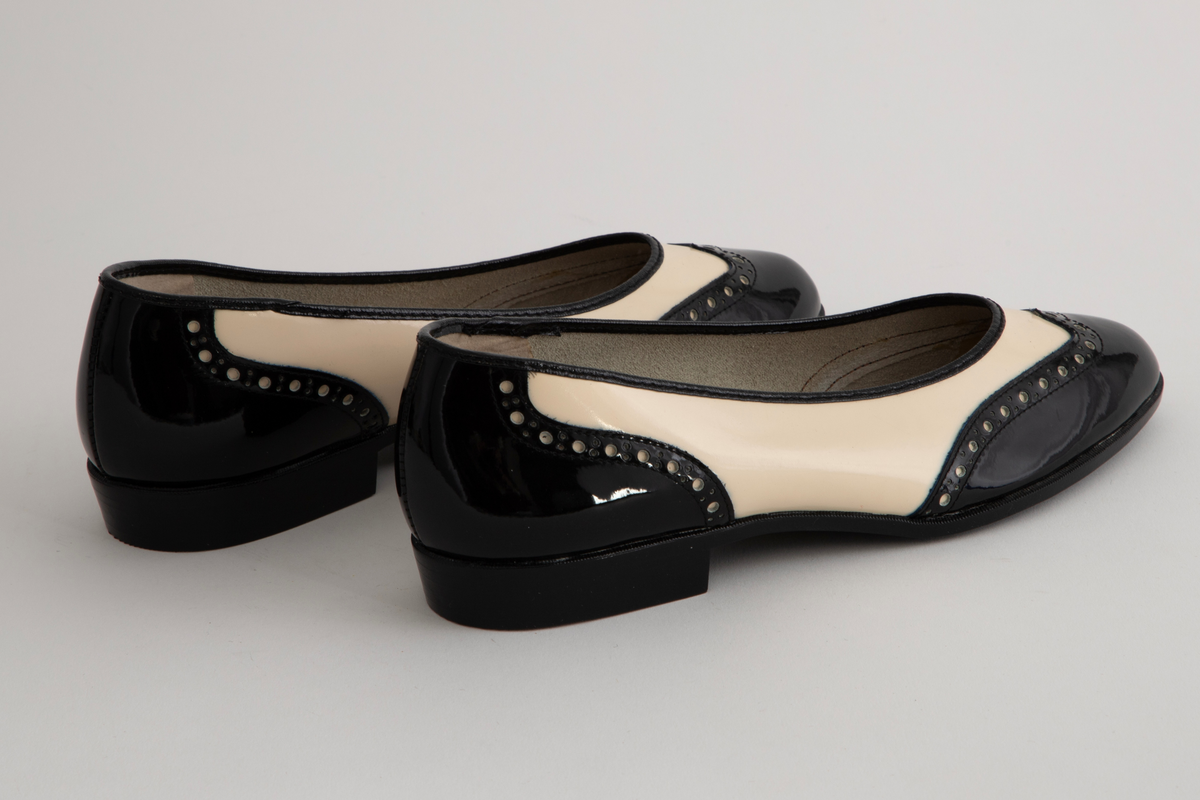 Ballerinasko/ pumps i svart- og hvittlakkert skinn med tilspisset, lett avrundet tå og bred, lav hæl. Tå- og hælkappe er svart med perforeringer, det øvrige hvitt. Svart kant rundt skoens åpning og svart hæl. Ufôret.