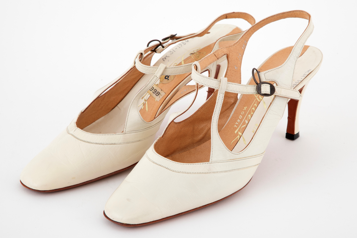 Høyhælte sko i tynt, hvitt skinn med tverr tå, åpen hæl og tynne remmer over vrist og ankel, lukket med spenne. Høy, tynn hæl. Lærsåle og innvendig fôr.