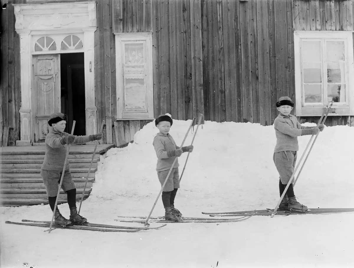 Ett gammalt fotografi som visar tre pojkar på skidor, vända åt höger, framför ett hus. Bilden är svartvit. 