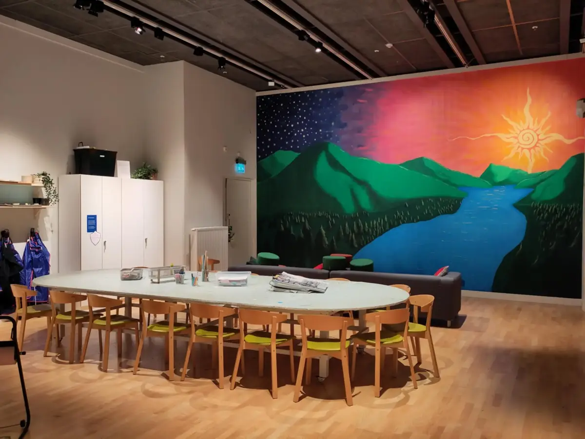 Bild på museets studio. Ett stort bord med stolar runt omkring och ett par skåp. På en av väggarna ser vi en stor väggmålning som visar ett landskap; gröna berg, skog och en älv som går mellan bergen. Himlen skiftar från sol och dag till natt och stjärnor. 