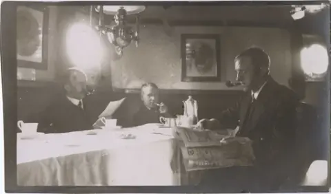 Tre menn i en skipssalong røyker pipe og leser aviser. Oskar Alf Gunvaldsen til høyre. Bilde av Kong Haakon VII og Kronprins Olav på veggen.