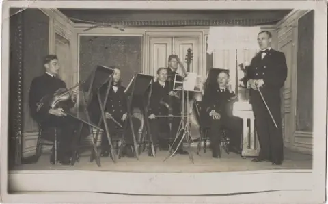 Fotopostkort. Orkester med fem mannlige musikere og en dirigent oppstilt med instrumenter og notestativ på en scene.