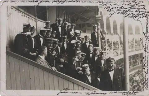 Fotopostkort. En rekke menn i skipsuniformer på en fullsatt tribune på ukjent sted. Oskar Alf Gunvaldsen sees i profil, sittende bak mann med flosshatt i front.