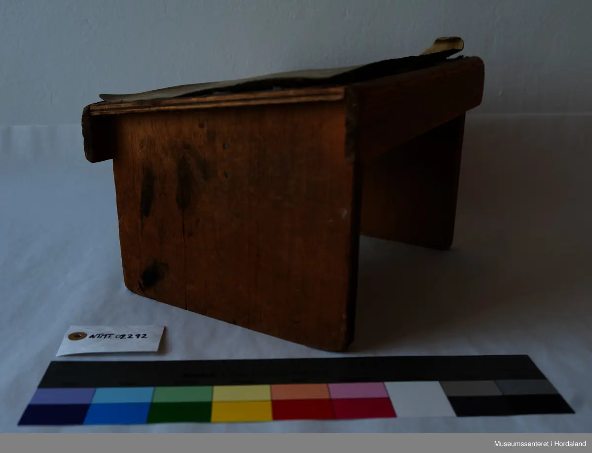 Liten, brun fotskammel av tre med linoleums-lignende lag på setet.