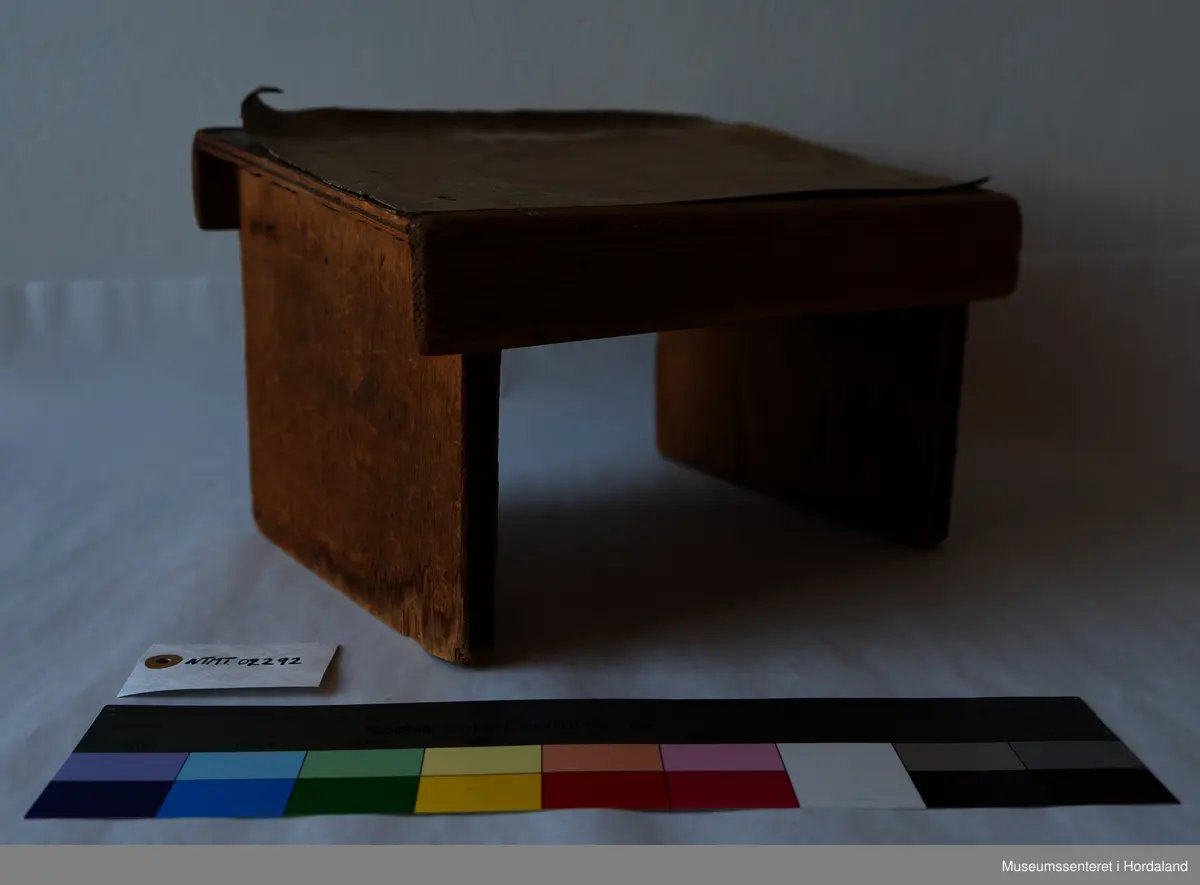 Liten, brun fotskammel av tre med linoleums-lignende lag på setet.