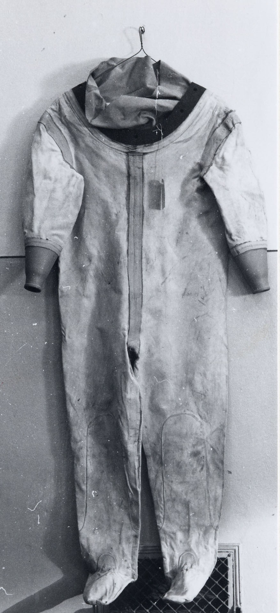 Tungdykardräkt av gummerad vit väv, i sömmarna förstärkt med gummerad beige väv. Hela fötter: i ärmarnas avslutning beige gummimanschetter. Halslinningen av svart gummi med hål för hjälmens fäste, samt innanför beige väv.

Skrivet med bläck i halslinningen datum för reparationer (1967 och 1982) samt signaturen "BH".