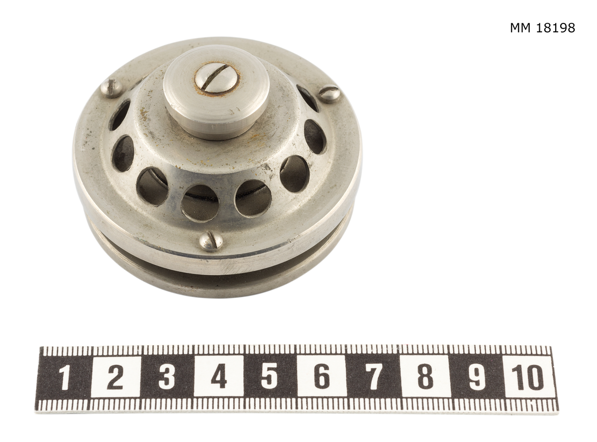 Andningsventil av stål bestående av två runda plattor: den översta med hål i mitten är gängad till den understa plattan som är försedd med ring av mindre hål på undersidan. Skruv i botten. Märkt med graverade bokstäver ifyllda med svart på ovansidan "Ventil till andningsapparat" samt "Minne av uppfinnaren Arne Zetterström ".