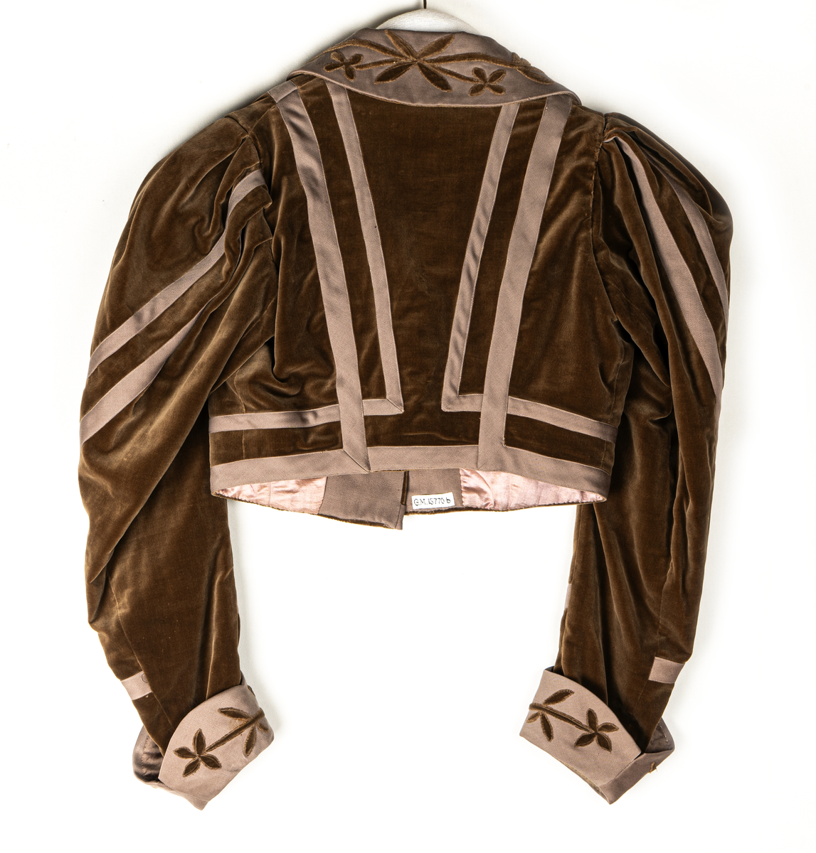 Del av dräkt/klänning.
b) Jacka, sammet. Krage och slag av gabardin med blommor av sammet. Knäppning fram. Långa puffärmar med slag. livet och ärmarna dekorerade med gabardinremsor. L fram 43cm, bak 37cm.