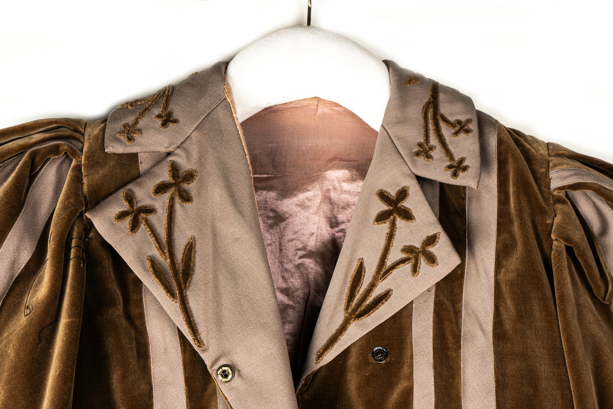 Del av dräkt/klänning.
b) Jacka, sammet. Krage och slag av gabardin med blommor av sammet. Knäppning fram. Långa puffärmar med slag. livet och ärmarna dekorerade med gabardinremsor. L fram 43cm, bak 37cm.