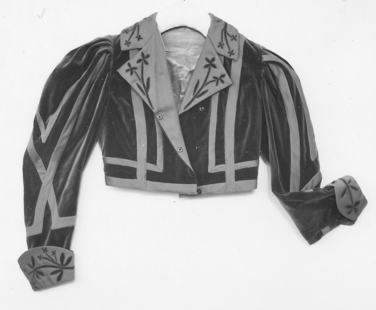 Del av dräkt/klänning.
b) Jacka, sammet. Krage och slag av gabardin med blommor av sammet. Knäppning fram. Långa puffärmar med slag. livet och ärmarna dekorerade med gabardinremsor. L fram 43cm, bak 37cm.