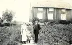 Bild av tre personer, en man, en kvinna och ett barn, som står utomhus framför en träbyggnad, troligen deras hem.
