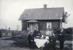 Bilden visar en grupp människor som sitter utomhus runt ett bord och fikar, bakom dem finns en stuga.