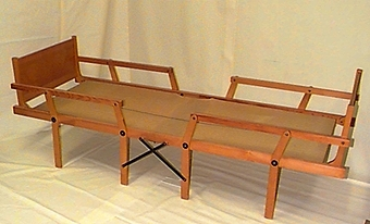 Rektangulär ram av lackerat trä. På båda kortsidorna sitter en rektangulär träskiva, höjd 287 mm, bredd 640 mm. Fyra ben utmed vardera långsida, fastsatta med skruv. Dessa ben är rörliga och sängen kan tryckas ihop, kortsida mot kortsida så att den ryms på liten yta. Sängbotten av brun juteväv med grön rand i mitten från kortsida till kortsida. I sängbotten på ena kortsidan sitter oval metall skylt med text "Wassbergs patent". Två svarta metallbågar under sängbotten och ett krysstag av svart metall på båda långsidornas mitt.