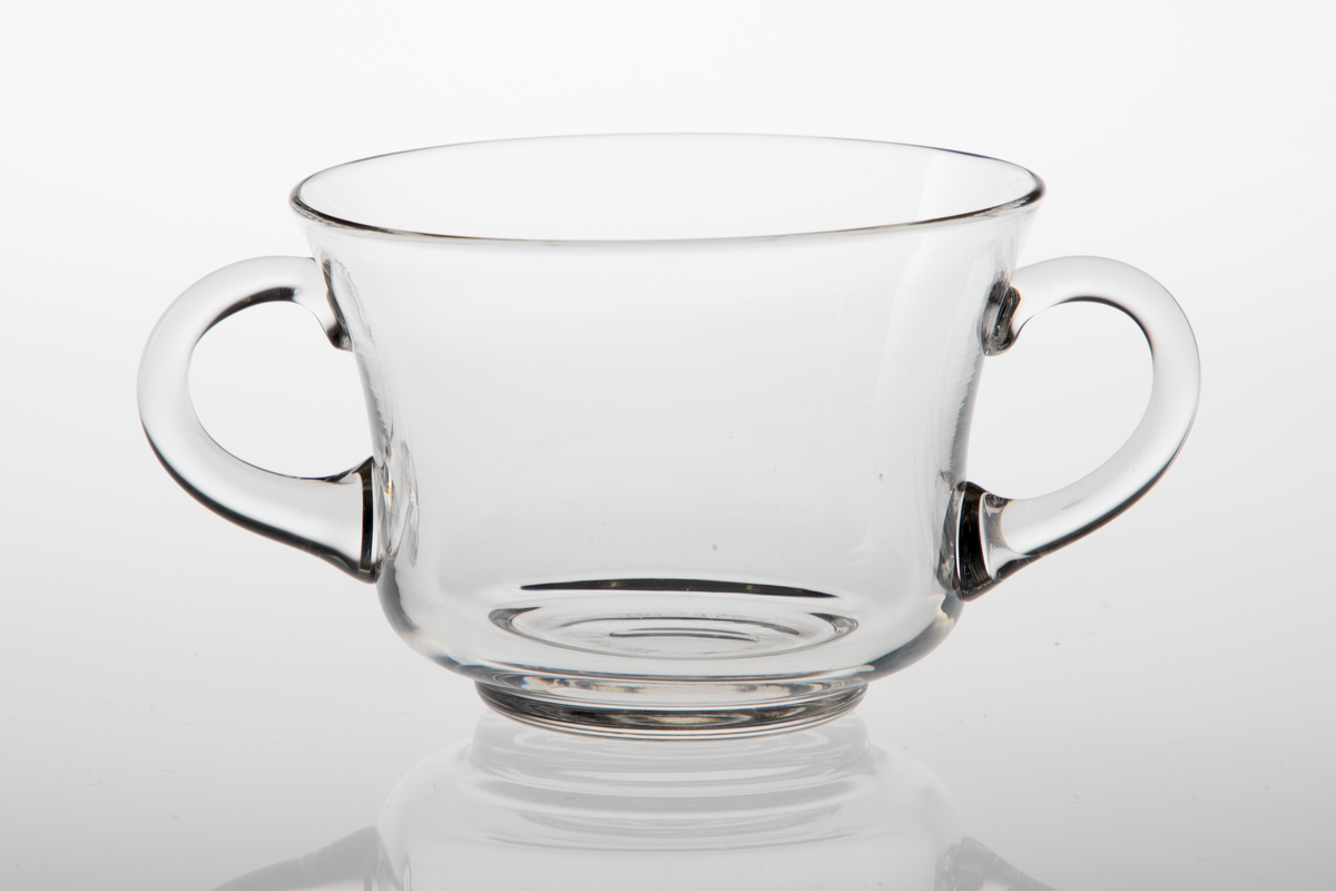 Sukkerkopp i klart glass. Konkav korpusform på lav fotring. To diametralt påsatte hanker.