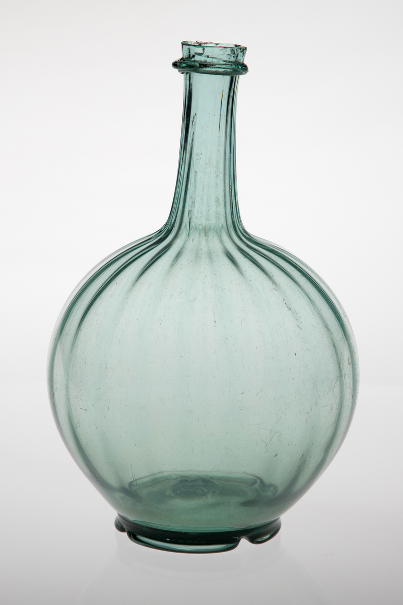 Kuleformet karaffel med lang hals. Glasset er lysegrønt, korpus og hals er dekket med vertikale rifler. Overgangen mellom hals og munning er markert med en smal ring. Korpus hviler på en tunget fotring med puntemerke.