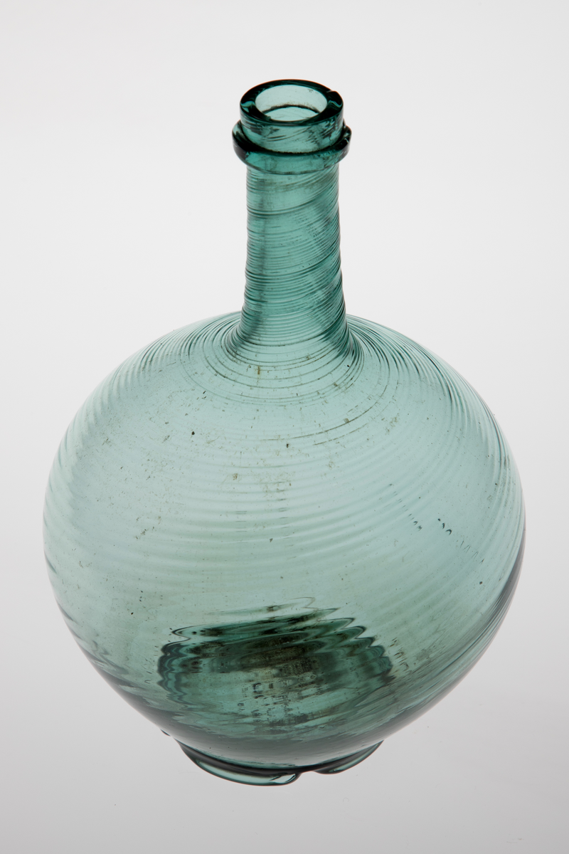 Kuleformet karaffel med lang hals. Glasset er lysegrønt, korpus og hals er dekket med tette, sterkt dreide rifler. Overgangen mellom hals og munning er markert med en smal ring. Korpus hviler på en tunget fotring med puntemerke.