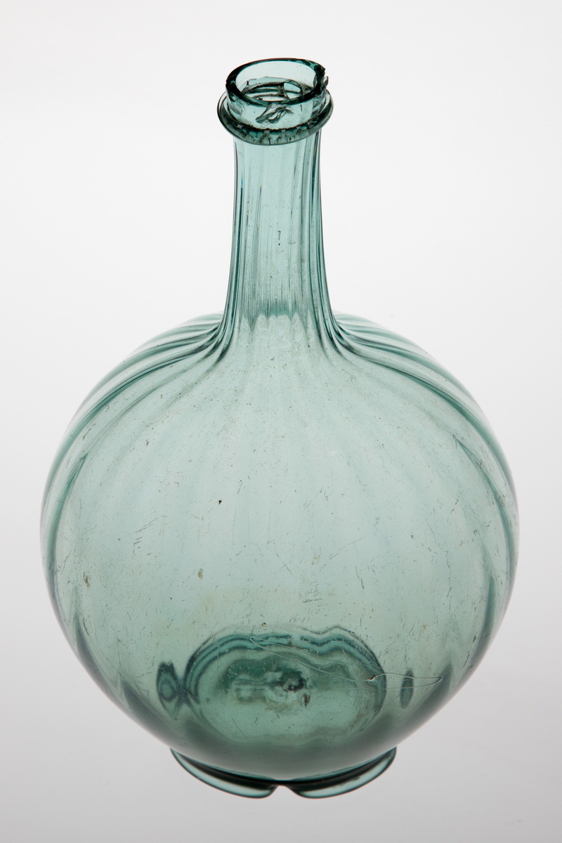 Kuleformet karaffel med lang hals. Glasset er lysegrønt, korpus og hals er dekket med vertikale rifler. Overgangen mellom hals og munning er markert med en smal ring. Korpus hviler på en tunget fotring med puntemerke.