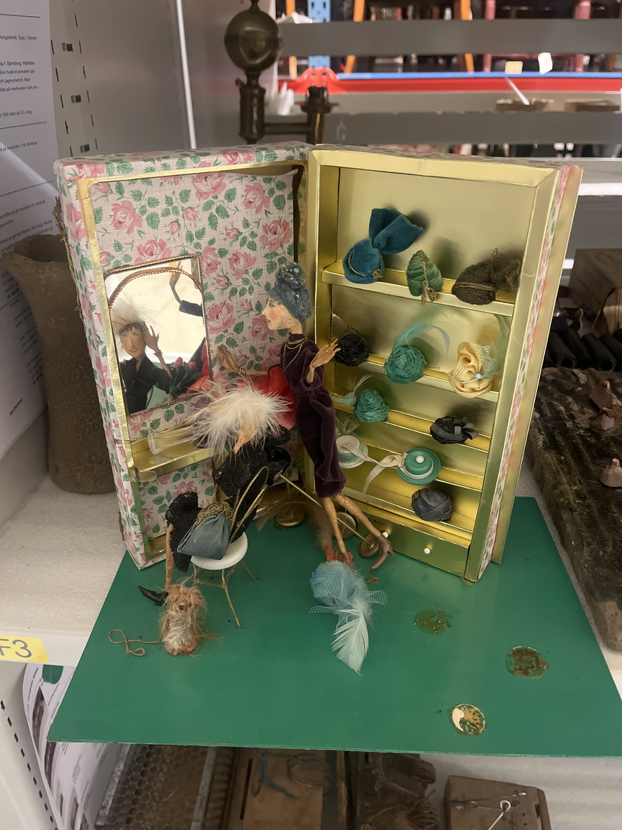 Ingår i serien "Små människor" tillverkade av Maja- Lisa Bengtsson i Borås mellan ca 1960-1980. Handgjorda dockor och scener av blandmaterial så som papier-maché, papper, och ståltråd med ansikten målade med vattenfärg.

Titel: Hattboutique