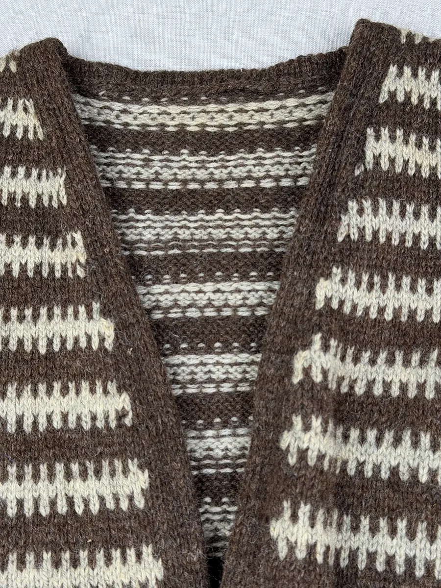 En v-ringad kofta, stickad i mönstrad slätstickning, av 3-trådigt ullgarn. Nederkanterna har en fåll, med ett avigt varv i vikningen, som är fastsydd med kaststygn.