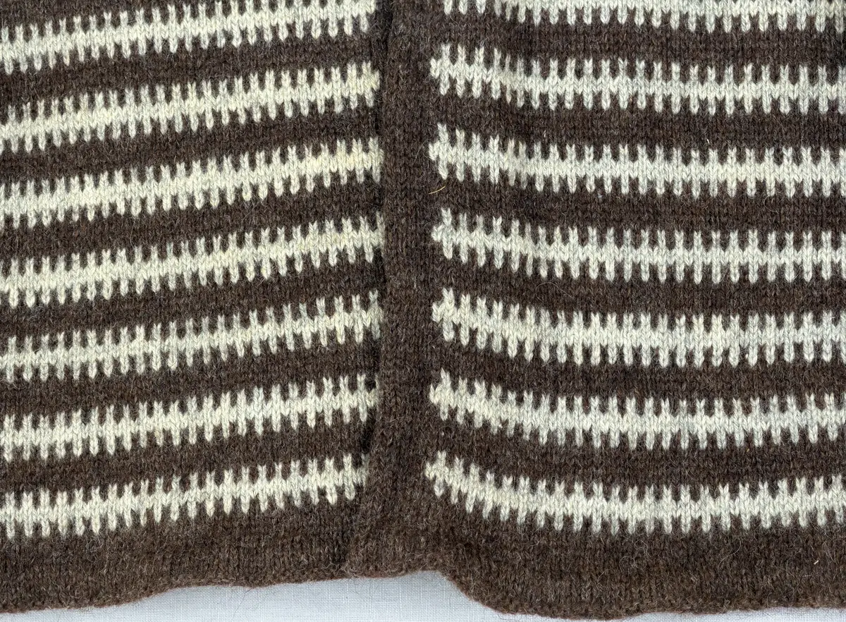 En v-ringad kofta, stickad i mönstrad slätstickning, av 3-trådigt ullgarn. Nederkanterna har en fåll, med ett avigt varv i vikningen, som är fastsydd med kaststygn.