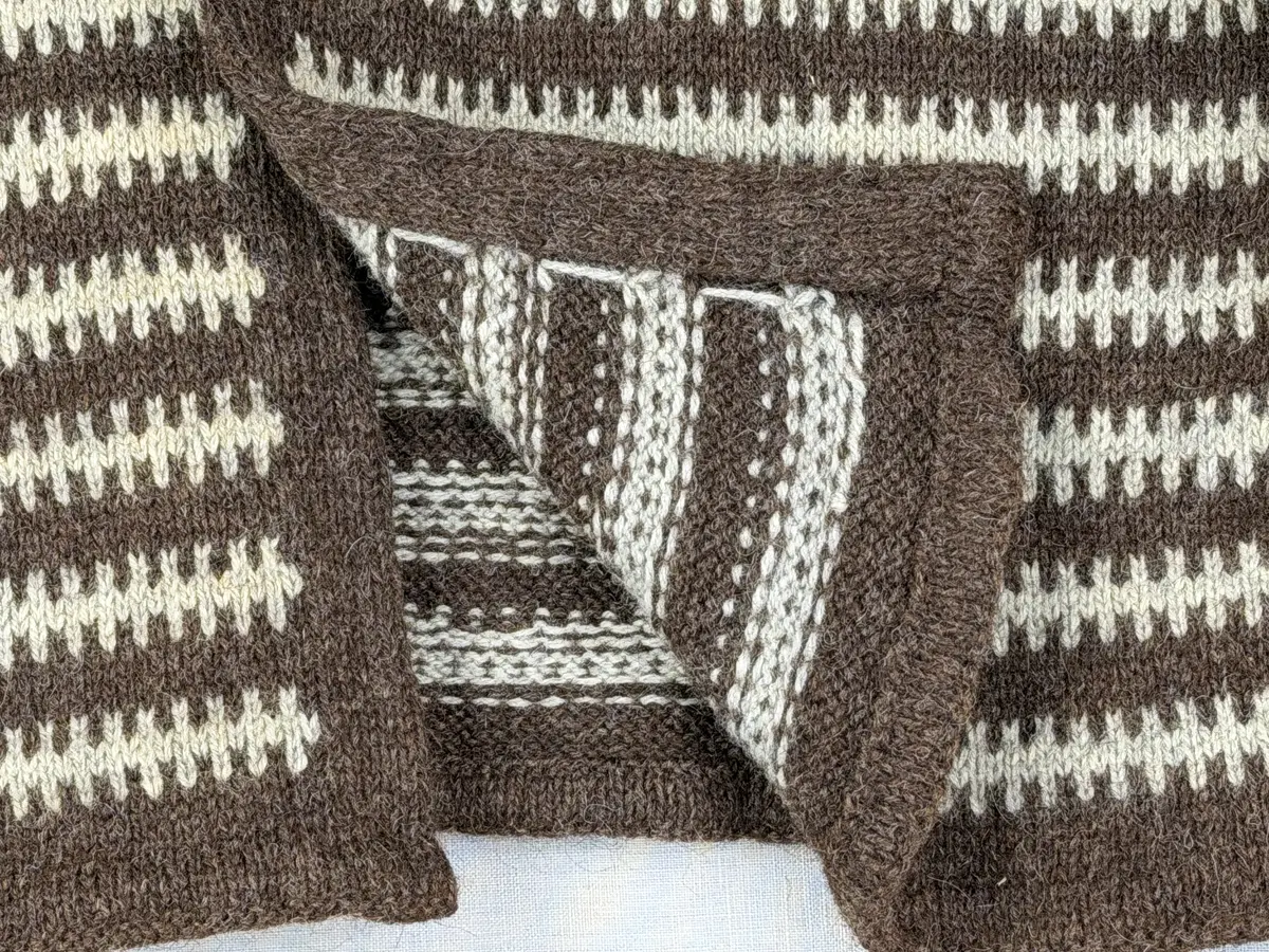 En v-ringad kofta, stickad i mönstrad slätstickning, av 3-trådigt ullgarn. Nederkanterna har en fåll, med ett avigt varv i vikningen, som är fastsydd med kaststygn.