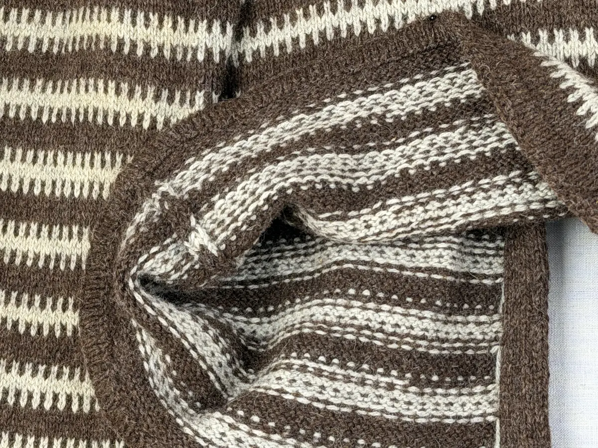 En v-ringad kofta, stickad i mönstrad slätstickning, av 3-trådigt ullgarn. Nederkanterna har en fåll, med ett avigt varv i vikningen, som är fastsydd med kaststygn.