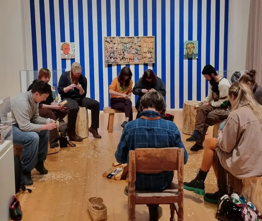 Personer sitter i en ring och täljer med en randig vägg med konstverk som bakgrund.