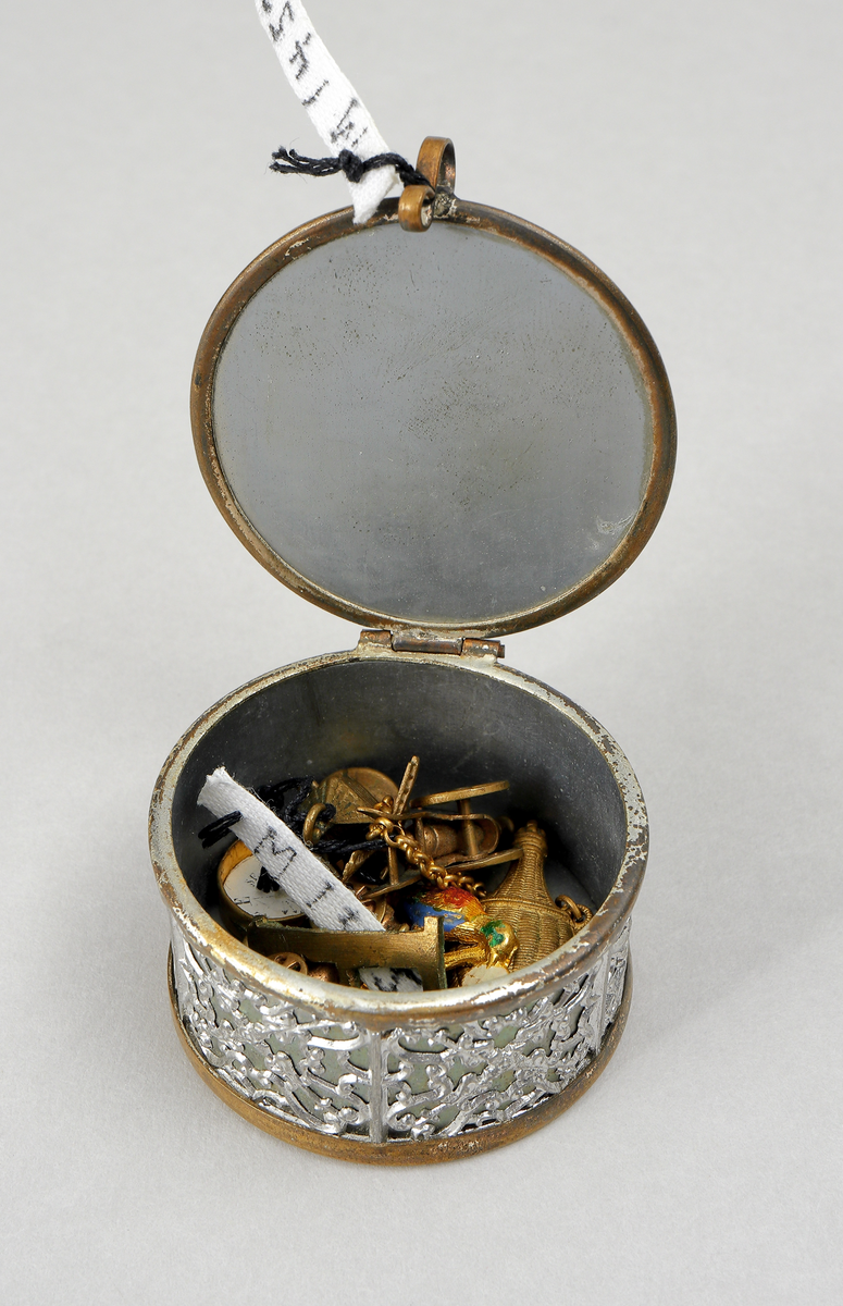 Nipperask innehållande diverse berlocker. Cylindrisk form med gångjärnförsett lock. Ornerat.