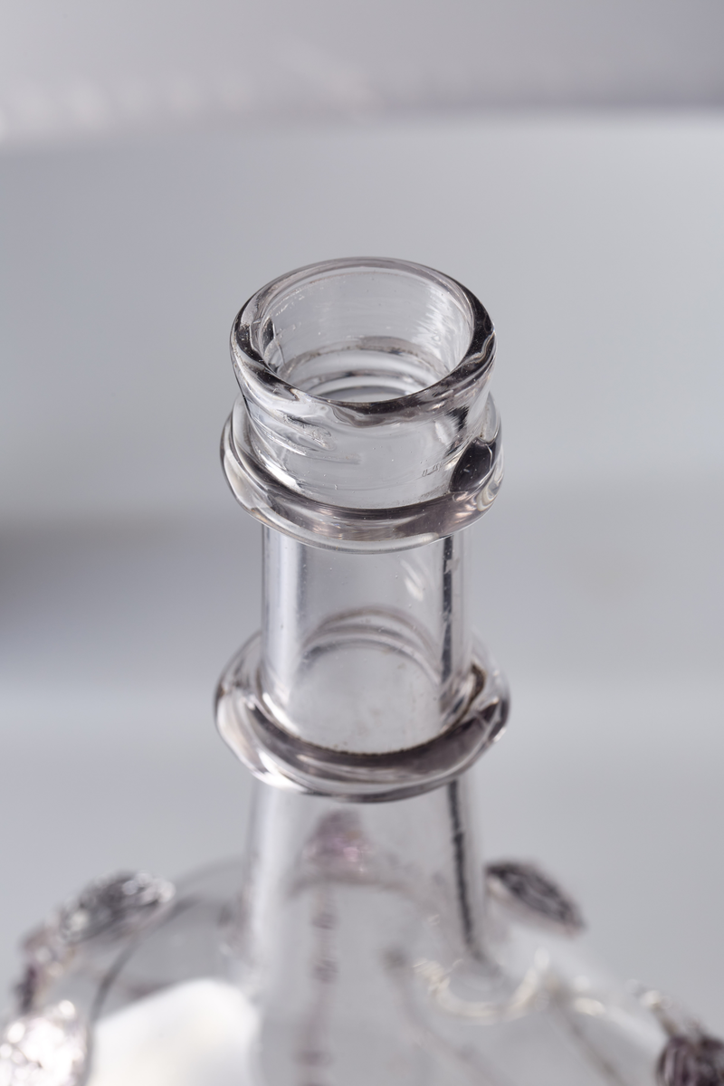 Åttekantet butelje med zirater og bringebær. Annenhver bringebær er svakt manganfarget. Pontmerke under flaska. To påsmeltede ringer på halsen.