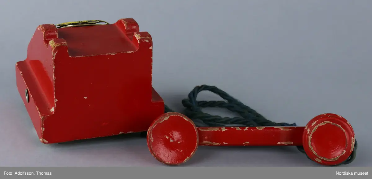 Leksakstelefon med nummerskiva och vidhängande telefonlur som sitter fast med en tvinnad lina av grönt garn. Etikett på framsidan med givarens telefonnummer textad med röd penna: "30272". På undersidan av telefonen finns en hålighet som ursprungligen kan ha innehållit en ringklocka. På undersidan är tre stöd av bakelit fastspikade, det fjärde saknas.