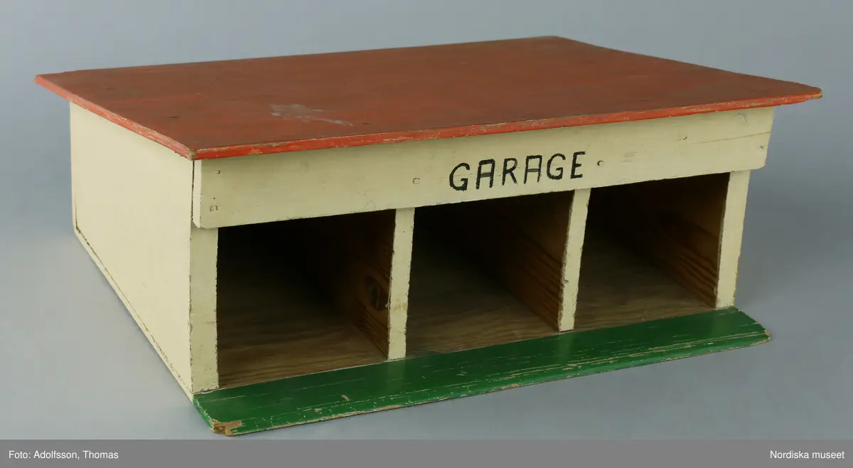 Garage av plywood med plats för tre bilar. Modellen är målad i ljusgult, med rött tak och grönt nedtill. Ordet GARAGE är skrivet i svart för hand, vilket kan tyda på att garaget kan vara hemsnickrat.