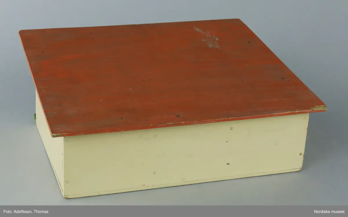 Garage av plywood med plats för tre bilar. Modellen är målad i ljusgult, med rött tak och grönt nedtill. Ordet GARAGE är skrivet i svart för hand, vilket kan tyda på att garaget kan vara hemsnickrat.