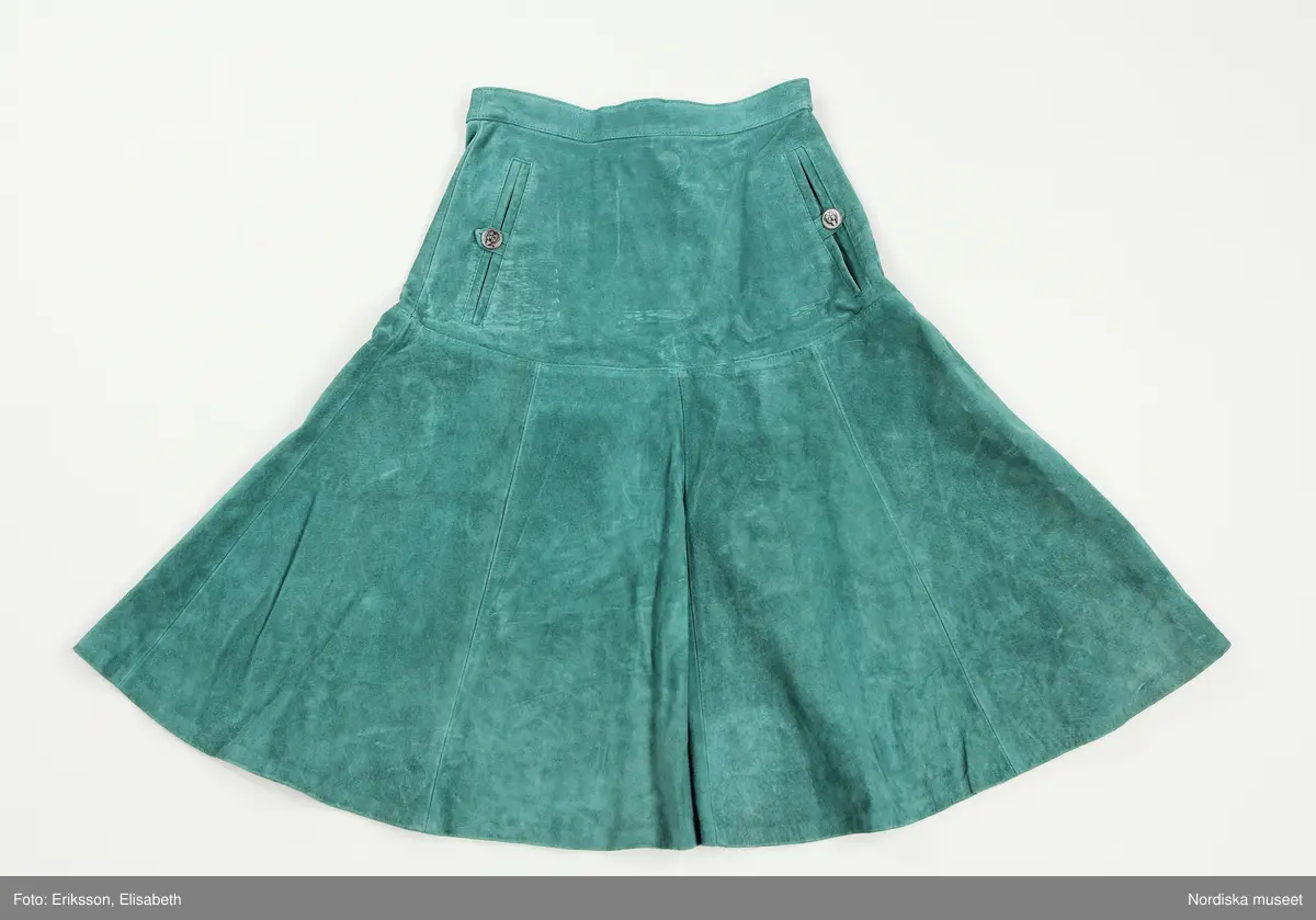 Grön mockakavaj och kjol. 
a) Dubbelknäppt kavaj som knäpps med metallknappar och passpoalerade knapphål. Knapparna är hela vägen upp (8 st på var sida) men två knappar döljs av slaget. Ett knapphål i midjehöjd och ett på slaget  på vänter sida, även de passpolerat och en grön plastknapp på insida att knäppa den med. Alla metallknapparna fastsydda med små "plastknappar" som förstärkning under, vid fickan är knapparna enbart fastsydda genom en lager av fickpåsarna.  
Axelklaffar som hålls på plats av metallknappar.
Söm mitt på ärmen med en stickning och en söm under ärmen.
Lodrätta infällda fickor med stolpe på framstycket 
Knytslelfar bak, fastsydda i längsgånde sömmar på bakstycket. 
På insidan av framstycket är det en maskinvävdetikett fastsydd. Texten säger 38 Tonnie 9-10 och runt detta står det skrivet i en cirkel MADE IN SWEDEN PRODUCED BY TONNIE DESIGN AB - STOCKHOLM -  SWEDEN - LW. Under cirkeln står det Von opp/xs

b) Kjol med besparing till stussen och utställda paneler 
På besparingen fram är det lodrätta passpolerade fickor med en knapp, rött foder i fickpåsarna. 
4 cm bred linning med dragkedja och knapp bak. 
På insidan stämplade nummer med vitt, samma nummer på alla delar. 

c) Underkjol, lös röd underkjol med två volanger sydda tillsammans på en besparing, den övre volangen lite kortare än den undre. Resår i midjan, trätt i en dragsko. 
På insidan av underkjolen är det en maskinvävdetikett fastsydd. Texten säger 38 Tonnie 9-10 och runt detta står det skrivet i en cirkel MADE IN SWEDEN PRODUCED BY TONNIE DESIGN AB - STOCKHOLM -  SWEDEN - LW. Under cirkeln står det Von opp/xs

/ Magdalena Fick 2025/07/10