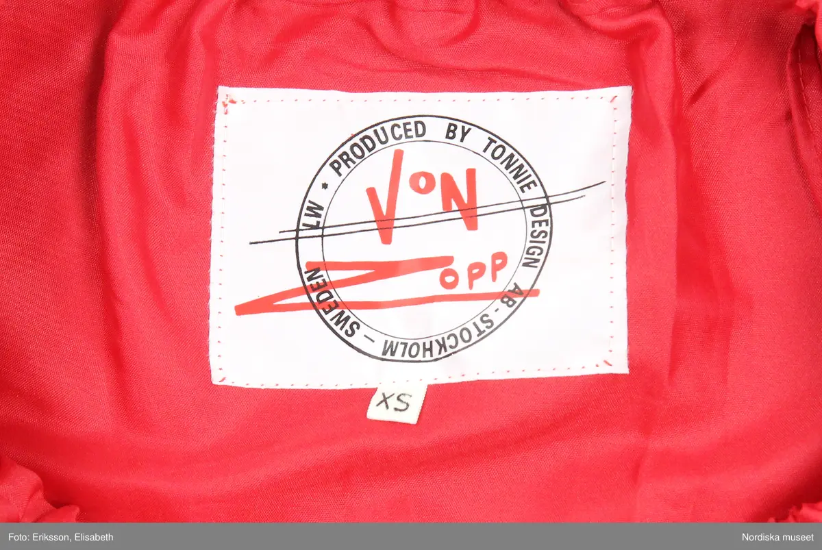 Grön mockakavaj och kjol. 
a) Dubbelknäppt kavaj som knäpps med metallknappar och passpoalerade knapphål. Knapparna är hela vägen upp (8 st på var sida) men två knappar döljs av slaget. Ett knapphål i midjehöjd och ett på slaget  på vänter sida, även de passpolerat och en grön plastknapp på insida att knäppa den med. Alla metallknapparna fastsydda med små "plastknappar" som förstärkning under, vid fickan är knapparna enbart fastsydda genom en lager av fickpåsarna.  
Axelklaffar som hålls på plats av metallknappar.
Söm mitt på ärmen med en stickning och en söm under ärmen.
Lodrätta infällda fickor med stolpe på framstycket 
Knytslelfar bak, fastsydda i längsgånde sömmar på bakstycket. 
På insidan av framstycket är det en maskinvävdetikett fastsydd. Texten säger 38 Tonnie 9-10 och runt detta står det skrivet i en cirkel MADE IN SWEDEN PRODUCED BY TONNIE DESIGN AB - STOCKHOLM -  SWEDEN - LW. Under cirkeln står det Von opp/xs

b) Kjol med besparing till stussen och utställda paneler 
På besparingen fram är det lodrätta passpolerade fickor med en knapp, rött foder i fickpåsarna. 
4 cm bred linning med dragkedja och knapp bak. 
På insidan stämplade nummer med vitt, samma nummer på alla delar. 

c) Underkjol, lös röd underkjol med två volanger sydda tillsammans på en besparing, den övre volangen lite kortare än den undre. Resår i midjan, trätt i en dragsko. 
På insidan av underkjolen är det en maskinvävdetikett fastsydd. Texten säger 38 Tonnie 9-10 och runt detta står det skrivet i en cirkel MADE IN SWEDEN PRODUCED BY TONNIE DESIGN AB - STOCKHOLM -  SWEDEN - LW. Under cirkeln står det Von opp/xs

/ Magdalena Fick 2025/07/10