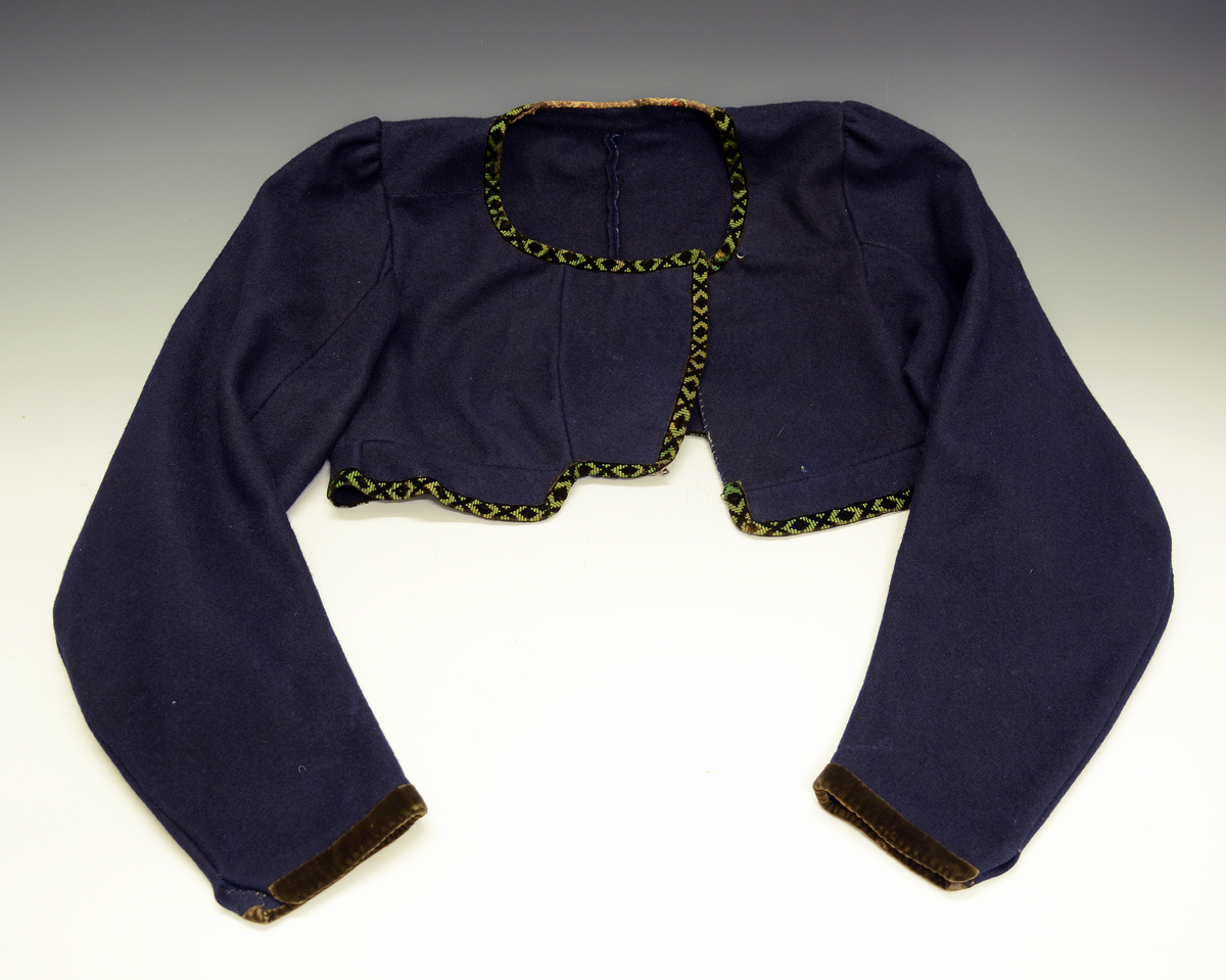 Drakt som består av A) overdel, B) stakk, C) trøye og D) forkle. Opplysninger fra protokollen: c) Trøye av blaatt vadmaal kanta med grøne og svartmynstra fløyelsborder (paa ermane svart fløyelskant). Trøyeblokune er skorne paa lag som uppluten, hev ei smal rispe nede bak, med 2 lask, og er til aa hekte framme. Dei tviblokka ermane hev litt rykkjing ved herdi og smaa stigle paa ermi framme. Sidd bak ikr. 29 cm
d) Bomullsfyrklæde med brogda fyrklædebunad, fyriklædet er graabrunt med strimur av svarte rankur fyldte med ljosare triblad. Sidd ikr. 1 m.