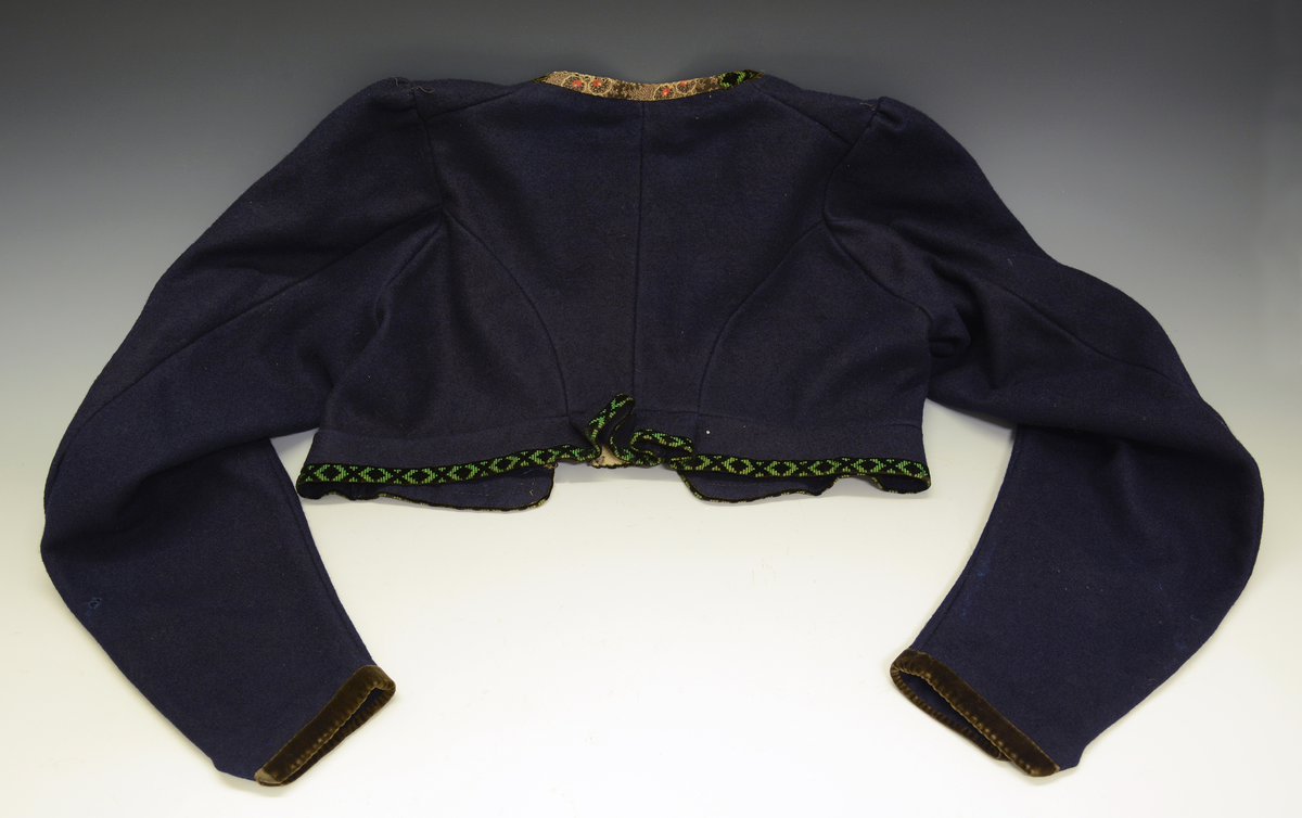 Drakt som består av A) overdel, B) stakk, C) trøye og D) forkle. Opplysninger fra protokollen: c) Trøye av blaatt vadmaal kanta med grøne og svartmynstra fløyelsborder (paa ermane svart fløyelskant). Trøyeblokune er skorne paa lag som uppluten, hev ei smal rispe nede bak, med 2 lask, og er til aa hekte framme. Dei tviblokka ermane hev litt rykkjing ved herdi og smaa stigle paa ermi framme. Sidd bak ikr. 29 cm
d) Bomullsfyrklæde med brogda fyrklædebunad, fyriklædet er graabrunt med strimur av svarte rankur fyldte med ljosare triblad. Sidd ikr. 1 m.