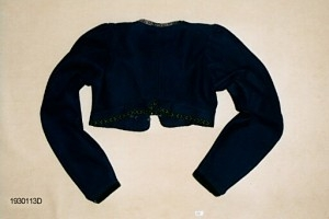 Drakt som består av A) overdel, B) stakk, C) trøye og D) forkle. Opplysninger fra protokollen: c) Trøye av blaatt vadmaal kanta med grøne og svartmynstra fløyelsborder (paa ermane svart fløyelskant). Trøyeblokune er skorne paa lag som uppluten, hev ei smal rispe nede bak, med 2 lask, og er til aa hekte framme. Dei tviblokka ermane hev litt rykkjing ved herdi og smaa stigle paa ermi framme. Sidd bak ikr. 29 cm
d) Bomullsfyrklæde med brogda fyrklædebunad, fyriklædet er graabrunt med strimur av svarte rankur fyldte med ljosare triblad. Sidd ikr. 1 m.