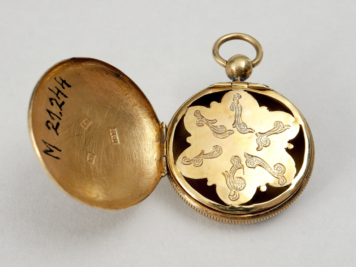 Medaljong av förgyllt silver. Utformad som en urboett. Runt kanterna stavdekor. Ytterboetterna varav den ena öppningsbar, är utvändigt graverade med med bladornament. Invändigt har den ett genombrutet inre boettlock försett med gångjärn. Gravyr i form av bladslingor. Medaljongen har förmodligen haft en s.k. luktsvamp placerad inuti. Medaljongen är helt förgylld in- och utvändigt. Stadstämpel F (möjligen Falkenberg). Mästarstämpel A.M, årtalsstämpel L5 (1865)
Inskrivet i huvudbok 1969.