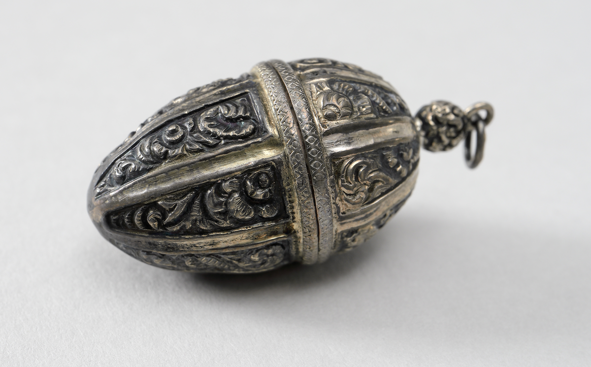 Hängsmycke i vitmetall utformat som ett ekollon med ornamentik. Gångjärnförsett lock. Insida förgylld, troligen har även smycket varit förgyllt utvändigt. I smycket går det att förvara piller eller dylikt.
Inskrivet i huvudbok 1969.