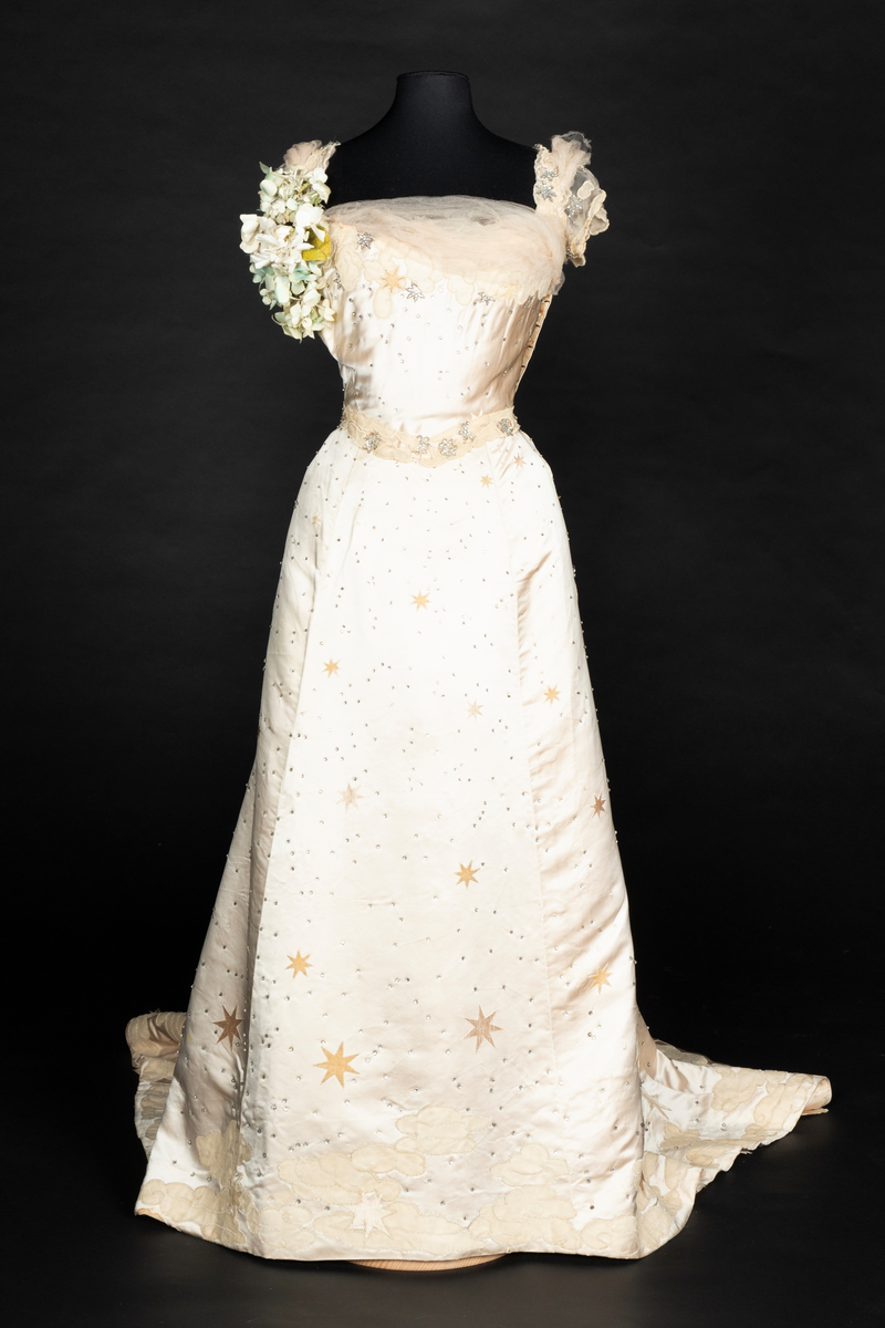 To-delt gallakjole med svært smal midje. Tettsittende, lavt ringet, kortermet livstykke og skjørt med slep. Fet, blank satengsilke, med ivevde stjerner i annen binding, deriblant stjerner av matt speilfløyel i en lys beige nyanse og skinnende blanke stjerner i en lysere nyanse. Kjolen er dekorert med små og store stjerner av begge typer, spredd utover stoffet, og i tillegg prydet med funklende "stjerner" av enkeltstående similistener/strass innfattet i blankt materiale. Kjolelivets halsringning, skulderpartier og midjeparti er også prydet med små stjerner av tettstilte similistener og noen innfelte stjerner av fløyel. Livstykkets utringning og nederkant, samt skjørtets fald og slep, er i tillegg prydet med applikerte, store og små skyer, konturert for hånd. De korte ermene og kjolelivets nederkant har kantborder av skyer sydd av chiffon, mens ringningen i tillegg er rikt dandert med bløt silketyll. Kjolelivet er avstivet med spiler, er fôret og har en innvendig lukning med snørehull midt bak. Denne er skjult av et ryggstykke som lukkes med skjulte hekter i venstre side. I tillegg har den et isydd midjebånd. Ringningen er dyp og rett skåret bak, mens fronten er asymmetrisk, med høyre skulder prydet av en stor og luftig bukett av Hortensia-blomster. Disse er utført i tekstilt materiale og delvis avstivet med metalltråd. 
Skjørtet er lett utskrådd, med vidden samlet midt bak, sluppet ut i et langt, rundet slep. Skjørtet er fôret, og har påsydd rynkekappe i nederkant av fôret. Denne er ikke faldet, men klippet i form av en tungekant.