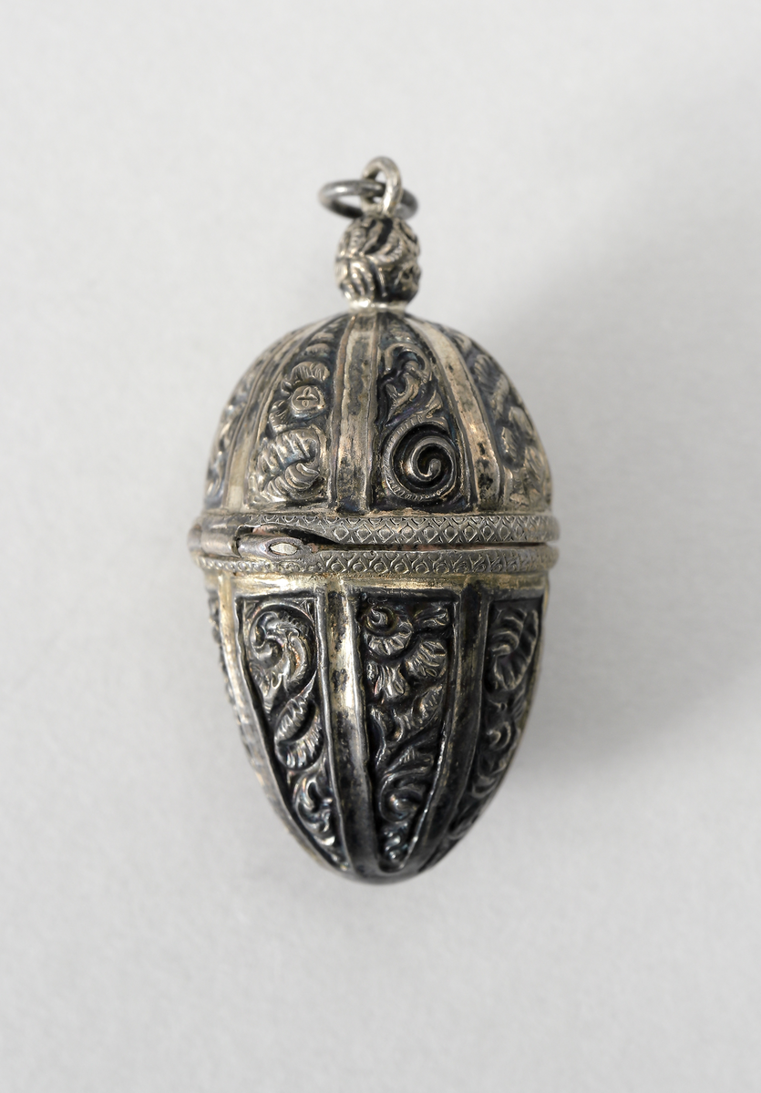 Hängsmycke i vitmetall utformat som ett ekollon med ornamentik. Gångjärnförsett lock. Insida förgylld, troligen har även smycket varit förgyllt utvändigt. I smycket går det att förvara piller eller dylikt.
Inskrivet i huvudbok 1969.