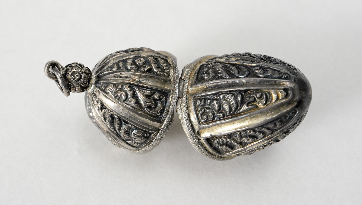 Hängsmycke i vitmetall utformat som ett ekollon med ornamentik. Gångjärnförsett lock. Insida förgylld, troligen har även smycket varit förgyllt utvändigt. I smycket går det att förvara piller eller dylikt.
Inskrivet i huvudbok 1969.