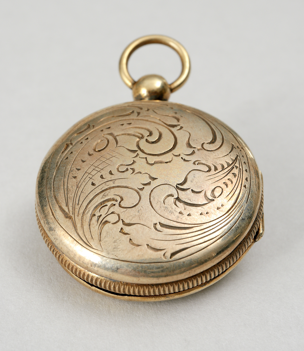 Medaljong av förgyllt silver. Utformad som en urboett. Runt kanterna stavdekor. Ytterboetterna varav den ena öppningsbar, är utvändigt graverade med med bladornament. Invändigt har den ett genombrutet inre boettlock försett med gångjärn. Gravyr i form av bladslingor. Medaljongen har förmodligen haft en s.k. luktsvamp placerad inuti. Medaljongen är helt förgylld in- och utvändigt. Stadstämpel F (möjligen Falkenberg). Mästarstämpel A.M, årtalsstämpel L5 (1865)
Inskrivet i huvudbok 1969.