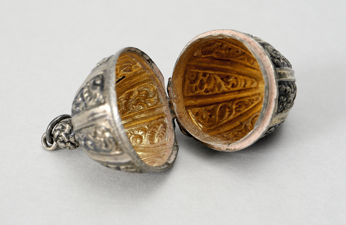 Hängsmycke i vitmetall utformat som ett ekollon med ornamentik. Gångjärnförsett lock. Insida förgylld, troligen har även smycket varit förgyllt utvändigt. I smycket går det att förvara piller eller dylikt.
Inskrivet i huvudbok 1969.