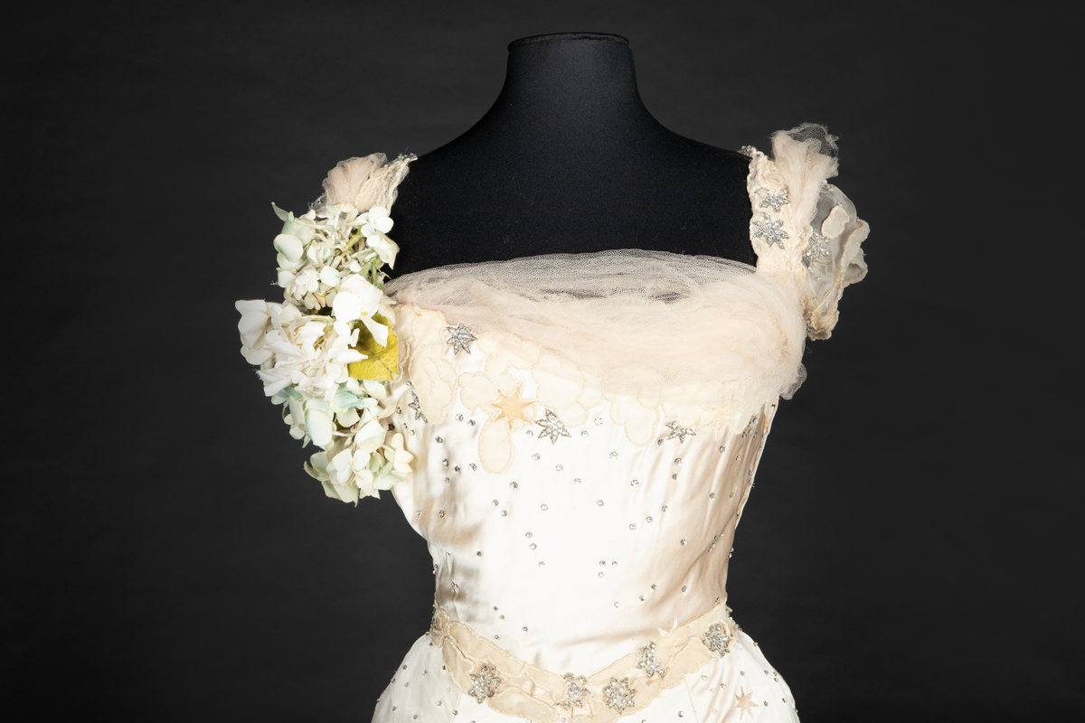 To-delt gallakjole med svært smal midje. Tettsittende, lavt ringet, kortermet livstykke og skjørt med slep. Fet, blank satengsilke, med ivevde stjerner i annen binding, deriblant stjerner av matt speilfløyel i en lys beige nyanse og skinnende blanke stjerner i en lysere nyanse. Kjolen er dekorert med små og store stjerner av begge typer, spredd utover stoffet, og i tillegg prydet med funklende "stjerner" av enkeltstående similistener/strass innfattet i blankt materiale. Kjolelivets halsringning, skulderpartier og midjeparti er også prydet med små stjerner av tettstilte similistener og noen innfelte stjerner av fløyel. Livstykkets utringning og nederkant, samt skjørtets fald og slep, er i tillegg prydet med applikerte, store og små skyer, konturert for hånd. De korte ermene og kjolelivets nederkant har kantborder av skyer sydd av chiffon, mens ringningen i tillegg er rikt dandert med bløt silketyll. Kjolelivet er avstivet med spiler, er fôret og har en innvendig lukning med snørehull midt bak. Denne er skjult av et ryggstykke som lukkes med skjulte hekter i venstre side. I tillegg har den et isydd midjebånd. Ringningen er dyp og rett skåret bak, mens fronten er asymmetrisk, med høyre skulder prydet av en stor og luftig bukett av Hortensia-blomster. Disse er utført i tekstilt materiale og delvis avstivet med metalltråd. 
Skjørtet er lett utskrådd, med vidden samlet midt bak, sluppet ut i et langt, rundet slep. Skjørtet er fôret, og har påsydd rynkekappe i nederkant av fôret. Denne er ikke faldet, men klippet i form av en tungekant.